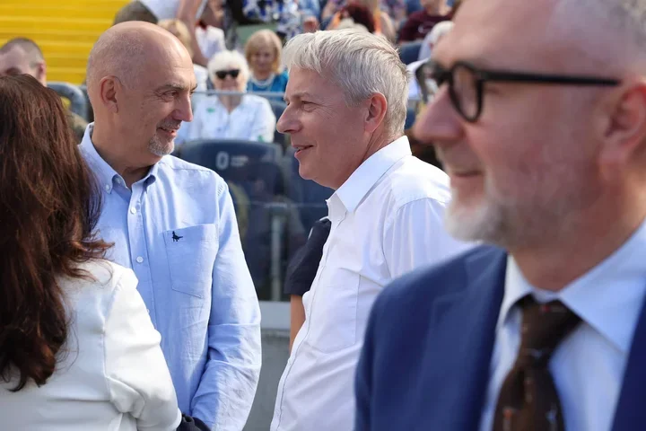 Trzech mężczyzn rozmawia na trybunach podczas plenerowego wydarzenia publicznego.