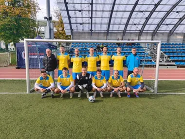 Drużyna piłkarska w żółto-niebieskich strojach pozuje do pamiątkowego zdjęcia przed bramką na stadionie z trybunami pod zadaszeniem.