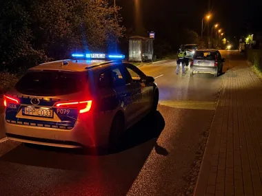 Policyjny radiowóz z włączonymi światłami nocnymi stoi na drodze za zatrzymanym samochodem osobowym, a dwóch funkcjonariuszy podchodzi do kierowcy.